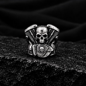 May include: A silver-toned metal ring featuring a detailed skull centered between two stylized engine cylinder blocks. The band includes a smaller circular emblem with a skull motif, showcasing a weathered, metallic finish with dark recessed details.