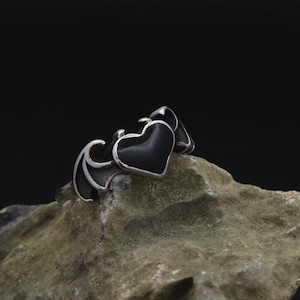 May include: A silver ring featuring a black heart-shaped centerpiece. The ring has a gothic design with wing-like accents on the sides of the heart. The ring is displayed on a textured, light brown rock against a dark background.