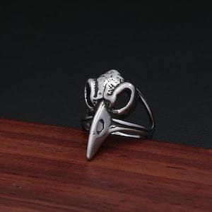 May include: A silver-colored ring featuring a detailed raven skull design. The ring has a pointed beak and a textured surface, with a dark background. The ring is made of metal and is a gothic-inspired accessory.
