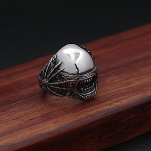 Peut inclure: Une bague argentée représentant une tête d'extraterrestre détaillée. La bague a un dessus lisse et réfléchissant et un visage texturé à bouche ouverte avec des dents pointues. La bague est posée sur une surface en bois.