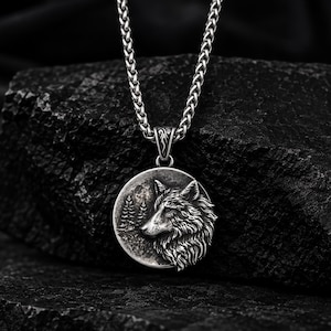 May include: A silver-toned metal necklace featuring a circular pendant with a detailed relief of a wolf head in profile, set against a background of pine trees and a moon. The pendant hangs from a thick, textured wheat chain with a decorative bail.