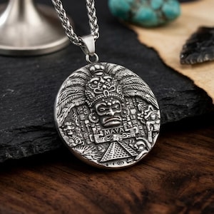 May include: A silver-toned circular pendant necklace featuring a detailed relief of a Mayan deity wearing an ornate headdress. The design includes a pyramid structure at the base and the word MAYA engraved in the center, suspended from a silver chain.