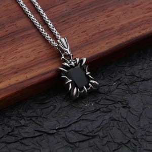 Pode incluir: Um colar com pingente prateado com uma pedra preta retangular. A pedra é colocada em uma moldura decorativa e pontiaguda. A corrente é de estilo tecido. O colar repousa sobre uma superfície de madeira escura.