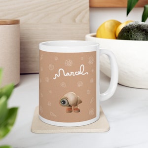 May include: White ceramic mug with a tan background featuring a cartoon seashell character and the name "Marcel" in white script. The mug is decorated with seashell illustrations.