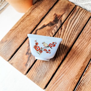 May include: A small, light blue ceramic cup with a bird and floral design. The bird is perched on a branch with red flowers. The cup is set on a wooden surface.