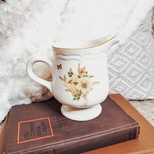 May include: White ceramic creamer with a floral design featuring yellow, brown, and green flowers. A small butterfly is painted on the side of the creamer.