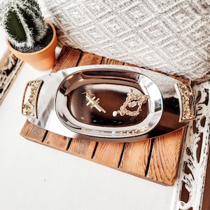 May include: A silver oval tray with gold filigree handles and accents, holding a gold cross earring and a gold necklace with a pearl. The tray sits on a wooden surface, with a small cactus in a terracotta pot in the background.
