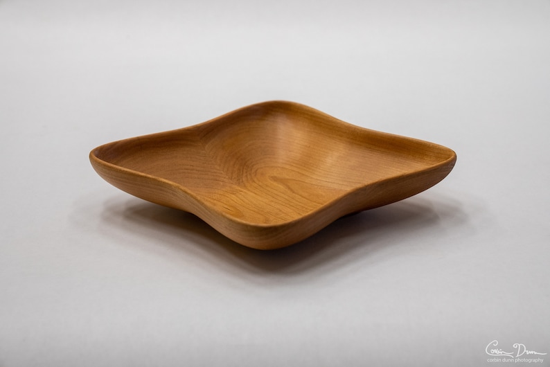May include: A square wooden bowl with a wavy, organic shape. The bowl is made of light brown wood and has a smooth, polished finish.