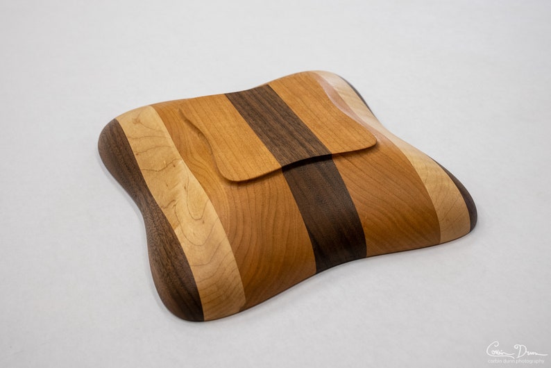 May include: A wooden bowl with a unique, wavy shape. The bowl is made from multiple types of wood, including walnut, maple, and cherry. The bowl has a lid that fits snugly inside.