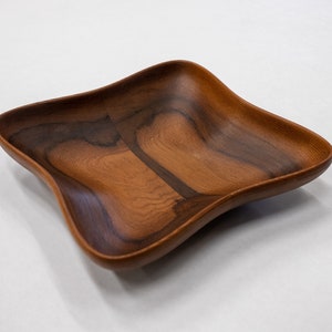 May include: A square wooden bowl with a dark brown wood grain pattern. The bowl has a slightly wavy edge and a deep, rounded interior.