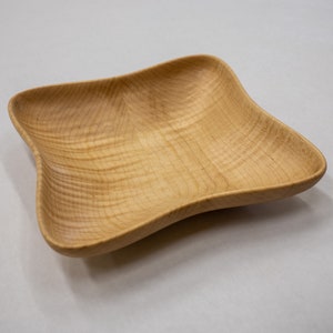 May include: A light wood square bowl with a slightly curved, rounded shape. The bowl is made of wood with a natural grain pattern.