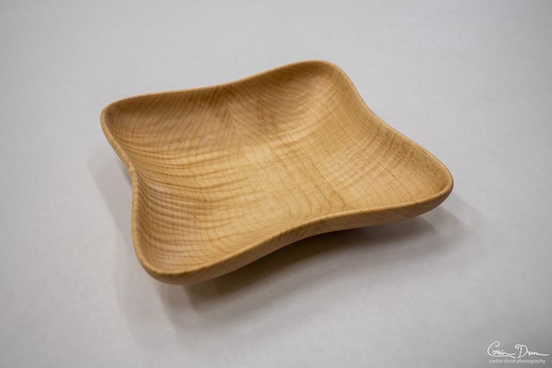 May include: A light wood square bowl with a slightly curved, rounded shape. The bowl is made of wood with a natural grain pattern.