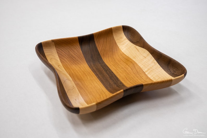 May include: A square wooden bowl with a wavy edge, made from different types of wood, including cherry, walnut, and maple. The bowl has a light brown, dark brown, and light wood grain pattern.