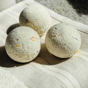 May include: Three spherical bath bombs, pale in colour, with yellow petal fragments, sit on a cream-coloured, striped towel. The bath bombs have a textured surface and appear handmade. The scene is lit with natural light, creating soft shadows.