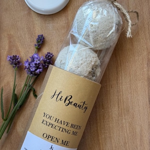 Op de afbeelding: Twee ronde, beige badbommen verpakt in een doorzichtige plastic hoes, vastgebonden met touw. De verpakking is voorzien van een beige label met de tekst Hi Beauty, YOU HAVE BEEN EXPECTING ME, OPEN ME en een lijst met ingrediënten.