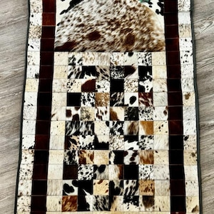 May include: A rectangular prayer rug made of cowhide. The rug features a patchwork design with squares of brown, black, and white cowhide. A dark brown border frames the rug, and a small prayer bead set is on the top.