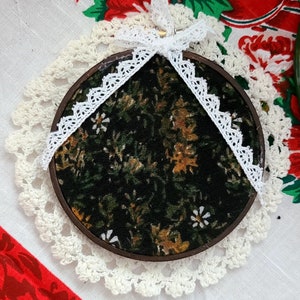 Peut inclure: Un cercle de broderie rond avec un motif floral en tissu dans des tons de vert, marron et blanc. Le cercle est décoré d'un ruban en dentelle blanche et repose sur un napperon en crochet blanc.