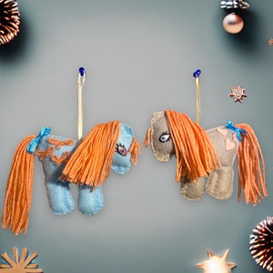 May include: Two felt horse ornaments with orange yarn hair, one is light blue and the other is light gray. Both have blue ribbon accents and are hanging from a string.