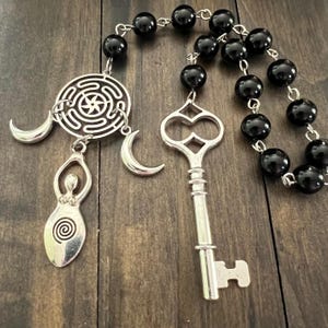 May include: A decorative hanging ornament featuring a string of smooth black beads connected to a silver-toned metal charm assembly. The charms include a circular labyrinth, two crescent moons, a stylized female figure, and a skeleton key.