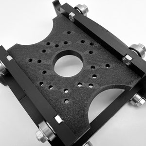 Puede incluir: Una placa de metal negro con un agujero circular en el centro y varios agujeros m&aacute;s peque&ntilde;os alrededor del borde. La placa est&aacute; unida a cuatro brazos de metal negro con rodamientos y tornillos.