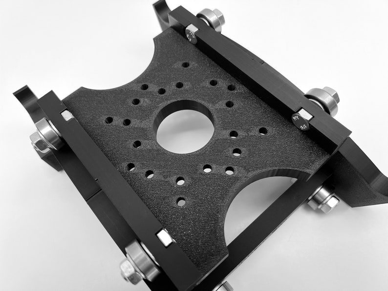 Puede incluir: Una placa de metal negro con un agujero circular en el centro y varios agujeros m&aacute;s peque&ntilde;os alrededor del borde. La placa est&aacute; unida a cuatro brazos de metal negro con rodamientos y tornillos.