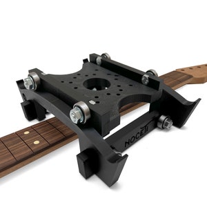 Puede incluir: Un jig de cuello de guitarra negro con un cuello de guitarra de madera sujeto en su lugar. El jig tiene una serie de rodillos y un logotipo NOCETI.