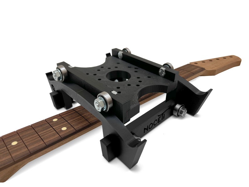 Puede incluir: Un jig de cuello de guitarra negro con un cuello de guitarra de madera sujeto en su lugar. El jig tiene una serie de rodillos y un logotipo NOCETI.