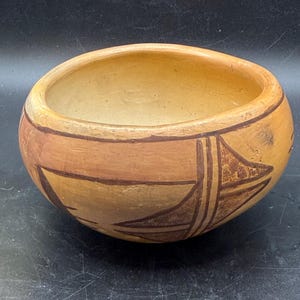 May include: A hand-painted, round ceramic bowl with a light brown interior and exterior. The exterior features geometric designs in shades of brown and tan. The bowl has a wide opening and a smooth, rounded shape, perfect for decorative use.
