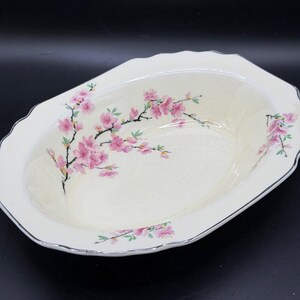 May include: Cream-colored oval serving dish with a scalloped edge and a silver trim. The dish is decorated with pink cherry blossom branches. A classic piece for serving food.
