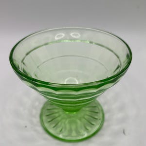 May include: A green glass dessert dish with a round bowl and a pedestal base. The bowl has horizontal ridges, and the base features a decorative pattern. The dish is made of transparent green glass.