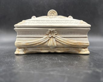 May include: A beige rectangular trinket box with a lid, featuring ornate floral and draped fabric details. The box has a decorative flower in the center and a small, round embellishment on the lid. The box is made of a light-colored material.