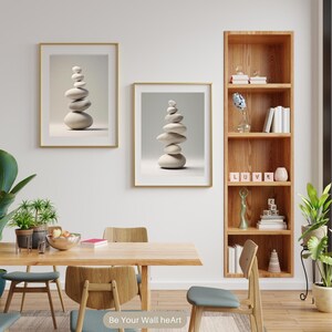 Puede incluir: Dos impresiones enmarcadas de piedras blancas apiladas sobre un fondo gris claro. Las impresiones est&aacute;n colgadas en una pared blanca sobre una mesa de comedor de madera con cuatro sillas. Una estanter&iacute;a de madera con estantes llenos de libros, objetos decorativos y un peque&ntilde;o jarr&oacute;n con flores azules se encuentra a la derecha de la imagen. El texto "Be Your Wall heArt" est&aacute; en la parte inferior de la imagen.