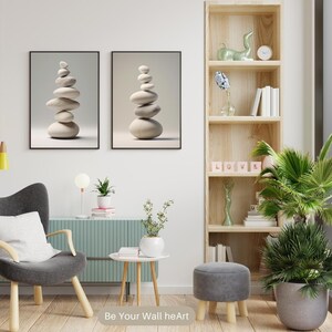 Puede incluir: Dos impresiones enmarcadas de piedras blancas apiladas sobre un fondo beige claro. Las impresiones est&aacute;n colgadas en una pared blanca sobre un sill&oacute;n gris con un coj&iacute;n blanco. Una estanter&iacute;a de madera con una escultura de cer&aacute;mica verde, libros y un reloj se encuentra a la derecha de la imagen. Una planta verde en maceta se encuentra en la esquina de la habitaci&oacute;n.
