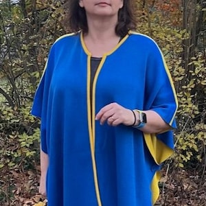 May include: A blue knitted poncho with yellow trim. The poncho has a long, flowing design and is open in the front. The model is wearing black pants.