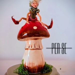 Peut inclure: Une sculpture de champignon en céramique avec un chapeau rouge et marron, un pied crème et une figurine de fée assise. La fée est vêtue de vert et de rouge, avec des cheveux blancs. La sculpture repose sur une base dorée avec de la mousse verte. Le texte "PEN SE" est visible.