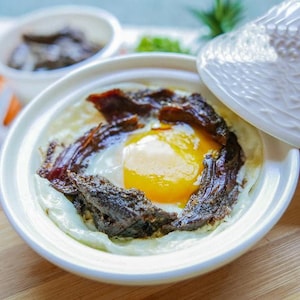 May include: A white ceramic dish with a lid, containing a fried egg with a runny yolk, topped with dark brown meat and a few pieces of dried meat.