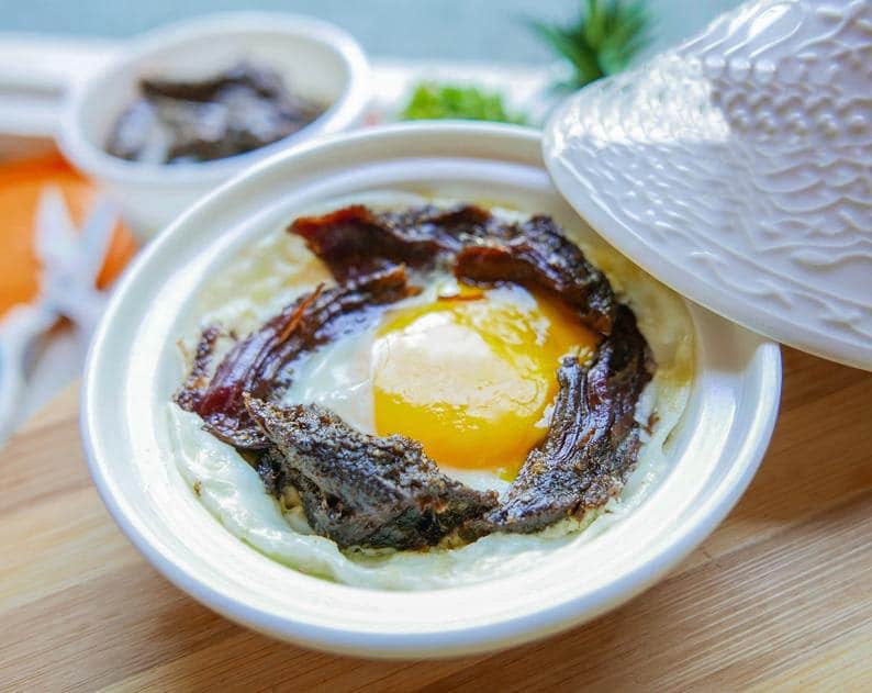 May include: A white ceramic dish with a lid, containing a fried egg with a runny yolk, topped with dark brown meat and a few pieces of dried meat.