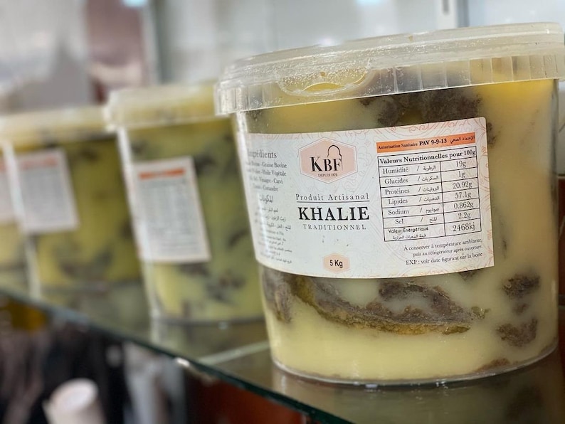 May include: A clear plastic container filled with a yellow and brown substance labeled "KHALIE TRADITIONNEL" and "Produit Artisanal". The container is on a shelf with other similar containers. The label also includes nutritional information in French and Arabic.