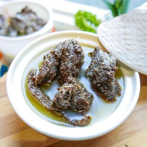 May include: A white ceramic bowl with a lid, filled with a dark brown stew or sauce. The stew is thick and has a glossy sheen. The bowl is sitting on a wooden surface.