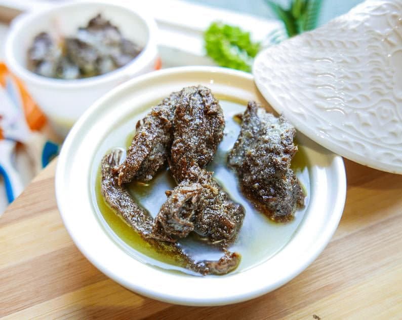 May include: A white ceramic bowl with a lid, filled with a dark brown stew or sauce. The stew is thick and has a glossy sheen. The bowl is sitting on a wooden surface.