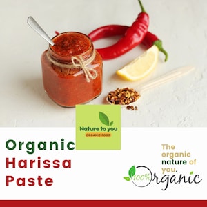 May include: A glass jar of red organic harissa paste with a brown twine bow. The jar is on a white surface with red chili peppers, a lemon wedge, and a spoon with red pepper flakes.