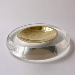 May include: A clear acrylic ashtray with a gold metal insert. The ashtray is round and has a slightly raised edge.