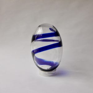 以下が含まれることがあります： 青い渦巻き模様の透明なガラスの花瓶。花瓶は楕円形で、白い台座の上に置かれています。