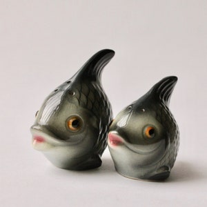 以下が含まれることがあります： 魚の形をしたセラミック製の塩コショウ入れのペア。魚はグレーで、黒と白のディテールが施され、黒目のオレンジ色の瞳をしています。魚はピンクの口をしていて、互いに向き合っています。