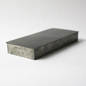 May include: Rectangular metal box with a dark gray lid and a weathered, industrial appearance. The box is made of a light gray metal and has a simple, minimalist design. The box is empty and sits on a white surface.