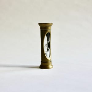 May include: A brass-coloured hourglass. The glass chamber holds white sand, and the design features a classic, cylindrical shape. The hourglass is a decorative item, suitable for a desk or shelf. The height is approximately 10 cm.