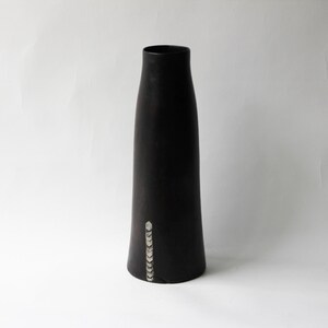 Peut inclure: Un grand vase noir avec une base légèrement évasée et un col étroit. Une ligne verticale de petites formes géométriques blanches orne la partie inférieure du vase. Le vase a une finition lisse et mate sur fond blanc.