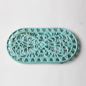 May include: A turquoise cast iron trivet with an oval shape and intricate openwork floral design. The trivet has a distressed finish, showing hints of rust, and is designed to protect surfaces from hot cookware.