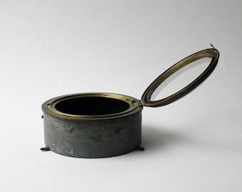 Könnte beinhalten: Ein zylindrischer Metallbehälter mit einem aufklappbaren, durchsichtigen Glasdeckel. Der Behälter hat ein dunkles, gealtertes Aussehen mit einem Messingrand und kleinen Füßen. Der Deckel ist geöffnet und gibt den Blick ins Innere frei.