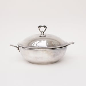 May include: A silver-colored serving dish with a lid and two side handles. The lid is topped with a heart-shaped handle. The dish has a rounded shape and is likely made of metal. The background is plain white.